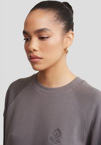 Szara bluza z okrągłym dekoltem, raglanowymi rękawami oraz subtelnym haftowanym logo na piersi. Gładka tekstura materiału, minimalistyczny design.