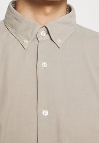 Gros plan sur une personne portant une chemise beige à boutons avec un col boutonné et des boutons blancs.
