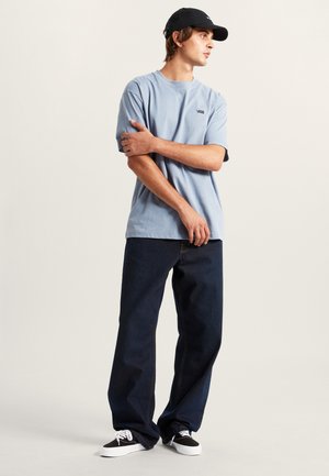 Giovane uomo con cappellino nero, t-shirt Vans azzurro chiaro, jeans scuri larghi e sneaker Vans nere, in piedi con le braccia incrociate.