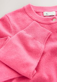 Lyserød strikket sweater med rund halsudskæring, ribstrikkede ærmekanter og en subtil tekstur, med en lille knapdetalje nær skulderen.