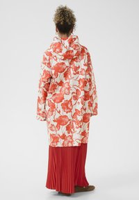 Kvetinový pršiplášť v krémovej a červenej farbe s kapucňou, dlhými rukávmi a voľným strihom; zdobený živým kvetinovým vzorom. Kombinuje sa so skladanou červenou sukňou.