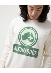 Sudadera de color crema con un gráfico verde de Australia, un signo más y el texto "ALTONADOCK" debajo. Textura suave y corte relajado.