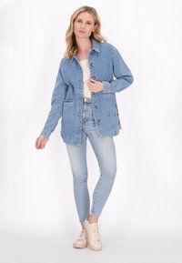 Vaaleansininen farkkutakki, jossa on nappikiinnitys, kaksi etutaskua ja rento leikkaus, yhdistettynä vaaleansinisiin kapeahelmaisiin farkkuihin ja beigeihin lenkkareihin.