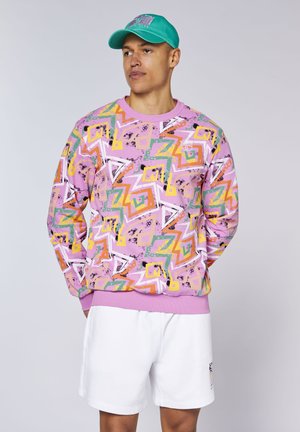 Mann mit türkisfarbener Kappe, pinkem Sweatshirt mit bunten geometrischen Mustern und weißen Shorts steht vor einfarbigem Hintergrund.