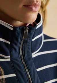 Tumesinine ja valge triibuline fliiskindel jakk kõrge kraega, millel on hõbedane tõmblukk ja kaubamärgi silt tõmbluku hoidjal. Pehme tekstuur.