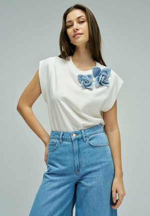 Mujer con cabello largo y castaño que lleva una blusa blanca sin mangas con aplicaciones de flores en denim azul y jeans de tiro alto azul claro, de pie con una mano en la cadera.