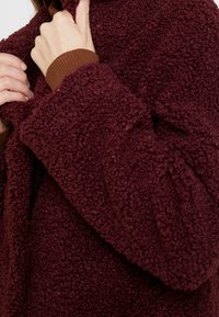 Giacca in fleece bordeaux con superficie morbida e testurizzata e polsini a costine marroni. Il tessuto è spesso e caldo, con una vestibilità ampia.