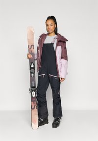 Suksen varusteet, joissa on vaaleanpunainen ja tummanlila takki, mustat haalarit vetoketjuilla ja mustat hiihtokengät. Pitelee suksea, jossa on vaaleanpunainen ja harmaa kuvio.