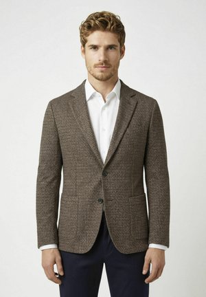 Mann mit lockigem Haar, der einen braunen strukturierten Blazer, ein weißes Hemd und dunkle Hose vor einem schlichten hellen Hintergrund trägt.