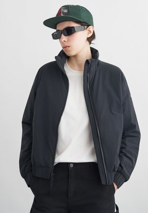 Tânăr purtând o jachetă neagră cu fermoar, cămașă albă, pantaloni negri, șapcă verde și ochelari de soare dreptunghiulari negri, stând cu mâinile în buzunare.