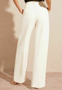 Wijde broek van lichtcrème stof, met een hoge taille, plooien en zijzakken, in combinatie met beige stilettohakken.