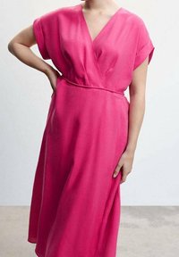 Femme portant une robe midi cache-cœur rose vif à manches courtes et col en V, debout avec une main sur la hanche.