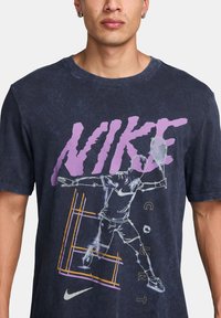 Camiseta de algodón azul marino con estampado en púrpura negrita de "NIKE", una gráfica de un jugador de tenis y un pequeño logo blanco de Nike en el dobladillo.