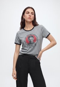 Femme aux longs cheveux foncés portant un t-shirt gris Stranger Things et un pantalon noir, debout avec une main sur la hanche sur un fond uni.