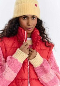 Czerwona pikowana kamizelka z wysokim kołnierzem; pod spodem różowy i kremowy sweter w wzory. Modelka ma na sobie żółtą prążkowaną czapkę. Widoczne kręcone włosy.