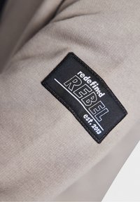 Beige fabric with a stitched black label; "redefined REBEL est. 2013" in white. The label has a rectangular shape and clear text.