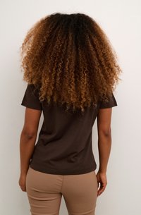 Camiseta corta de manga corta en color marrón, hecha de una tela suave, con una silueta ligeramente ajustada, combinada con pantalones ajustados beige, mostrando un peinado texturizado y rizado.