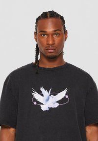 Camiseta de algodón negra de corte holgado que presenta un gráfico de dos palomas de color azul claro con acentos morados, centrado en el pecho.
