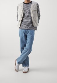 Gilet grigio chiaro con molte tasche sopra una maglietta a strisce bianco e nera a maniche lunghe, abbinato a jeans azzurri e scarpe bianche.
