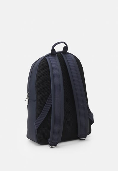 Sac à dos bleu marine avec un extérieur lisse, des panneaux en maille, des bretelles rembourrées et une poignée supérieure. Présente un zip argenté détaillé.