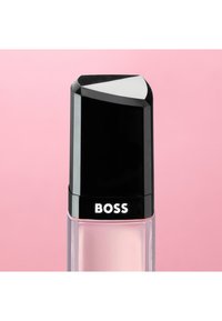 Flacon de parfum en verre transparent avec un bouchon noir, contenant un liquide rose pâle, étiqueté "BOSS", posé sur un fond rose uni.
