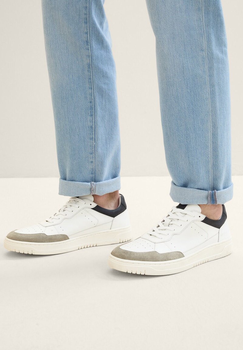 Weiße Sneaker mit grauen Wildlederakzenten und schwarzer Ferse, ausgestattet mit Perforationen und flachen Schnürsenkeln. Getragen mit hochgekrempelten hellblauen Jeans.