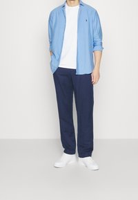 Hellblau gestreiftes Langarmshirt über einem weißen T-Shirt, kombiniert mit marineblauen Hosen und weißen Sneakers. Einfaches Design, lässige Passform.