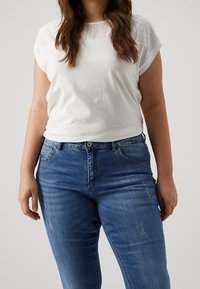 Vit bomulls t-shirt med ögla-detaljer vid axlarna, tillsammans med blå denimjeans med en klassisk passform och synliga sömdekorationer.