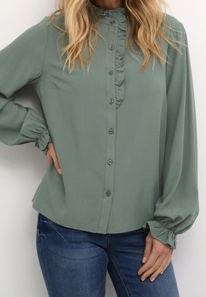 Vrouw draagt een lang mouwen blouse in saliegroen met een ruches kraag en manchetten, gecombineerd met blauwe spijkerbroek, hand op de heup.