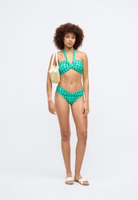 Zaļš rakstains bikini ar pagriežamu bandeau augšdaļu un augstā jostasvietā izgrieztiem apakšiem. Modelis tur nelielu salmu rokassomu un valkā gaismas sandales.