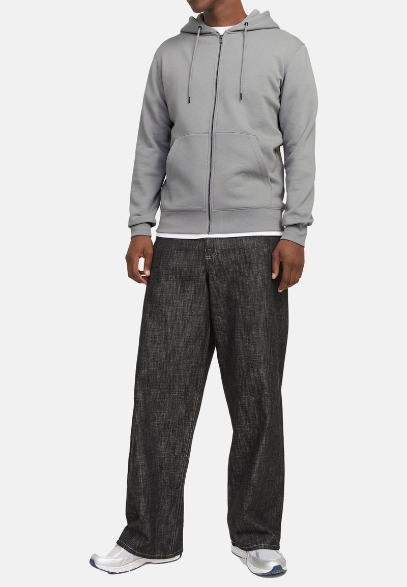 Grå zip-up hættejakke med en frontlomme, parret med løse, mørkegrå bukser med et diskret mønster. Hvid undertrøje og grå sneakers.