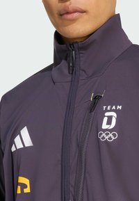 Ciemnofioletowa sportowa kurtka z wysokim kołnierzem, zapięciem na zamek, białymi i żółtymi akcentami logo oraz dopasowanym krojem.