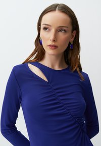 Langärmliges, tiefblaues Oberteil mit seitlichem Raffung und Ausschnittdesign. Begleitet von blau-goldenen Ohrringen mit runden Formen.
