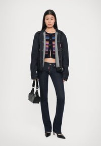 Veste en denim foncé avec une fermeture à boutons, haut en tricot court multicolore, jean foncé ajusté et chaussures à talons noires, tenant un sac à main noir.