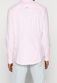 Camicia a maniche lunghe rosa chiaro realizzata in tessuto soffice, con colletto botton-down, yoke posteriore e orlo arrotondato. Accenti a bottoni visibili sulle maniche.