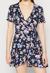 Robe portefeuille fleurie bleu marine, avec manches courtes, col en V profond et ceinture à nouer à la taille. Ornée de fleurs colorées et d'un ourlet à volants.