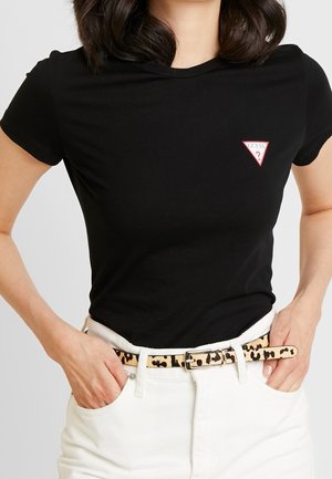 Kvinde iført en sort t-shirt med korte ærmer med et lille trekantet Guess-logo og hvide bukser med et beige leopardmønster bælte.