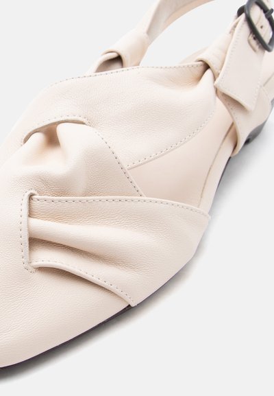 Μπεζ δερμάτινο σανδάλι με επίπεδη σόλα, με πιεσμένο μπροστινό λουράκι και ρυθμιζόμενο λουράκι στον αστράγαλο με μαύρη αγκράφα, σε λευκό φόντο.