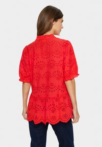 Rdeča bombažna bluzica z izrezovanjem, kratkimi zadrgami, valovitim robom in oprijetim pasom. Ima dekorativne izreze v cvetličnem vzorcu.