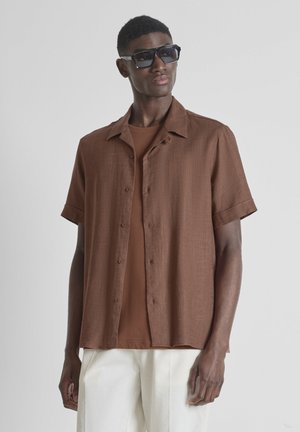 Man draagt een donkere zonnebril, een bruin overhemd met korte mouwen over een bruin T-shirt en witte broek, poserend tegen een lichte achtergrond.