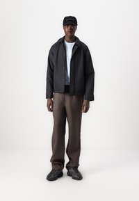 Black padded jacket, light blue t-shirt, brown trousers, and black sneakers. Man stands straight, with a black cap featuring white text.