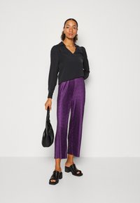 Blusa nera a maniche lunghe con scollatura a V, abbinata a pantaloni viola plissettati e scarpe nero con tacco blocco slip-on; tiene una borsa nera.