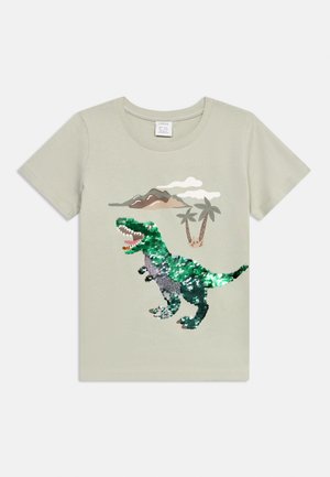 T-shirt beige à manches courtes avec un dinosaure en sequins verts et argentés, montagnes, palmiers et nuage imprimés sur le devant.