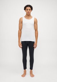 Weißes ärmelloses Tanktop und enganliegende schwarze Leggings, beide mit glatter Textur und simplem Design, getragen von einem stehenden Modell.