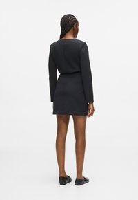 Donna con i capelli intrecciati, che indossa un abito nero aderente a maniche lunghe e scarpe basse nere, in piedi di spalle su uno sfondo bianco.