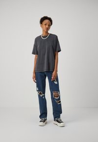 Dunkelgraues, übergroßes T-Shirt mit kurzen Ärmeln, kombiniert mit zerrissenen blauen Jeans mit großen Löchern. Schwarze und weiße Sneaker runden den Look ab.