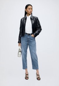 Frau trägt schwarze Lederjacke, weißes Hemd mit Knöpfen, blaue Jeans, schwarze Absätze und hält eine kleine weiße Handtasche vor einem einfarbigen Hintergrund.