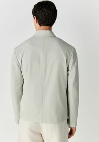 Chaqueta gris claro hecha de un tejido texturizado, con solapa en pico y un diseño limpio y recto, con detalles de costura sutiles en la parte posterior.