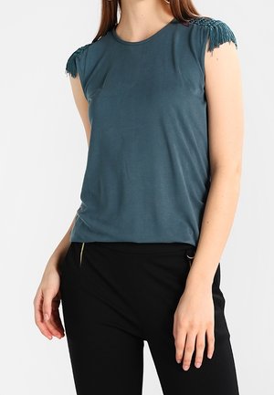 Teal short-sleeve top with a round neckline, featuring textured fringe accents on the shoulders. Smooth fabric and fitted shape.