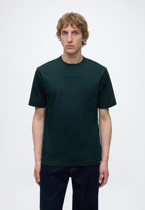 Jonge man met krullend blond haar, die een donkergroen T-shirt met korte mouwen en donkerblauwe spijkerbroek draagt, staand tegen een effen witte achtergrond.